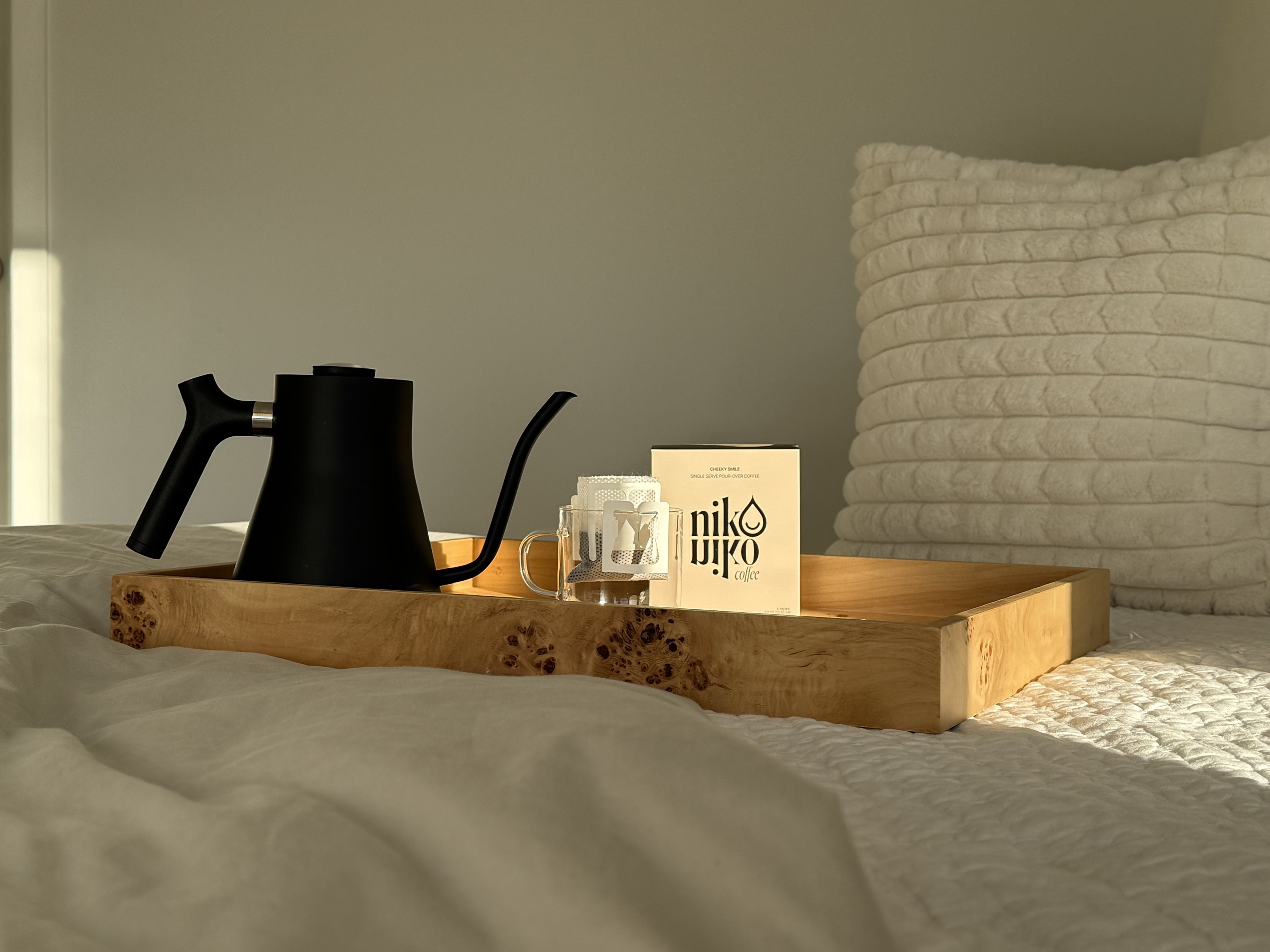 alt='Wooden tray with drip black coffee maker, glass mug, and coffee filter, niko niko coffee on a bed.' />.